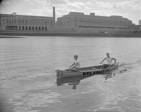 Two men paddle a canoe on a river with buildings on an island in the background.